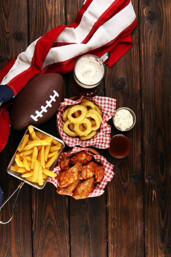A collection of onion rings, french fries and chicken wings in separate bowls on a wooden table next to a football and American flag.