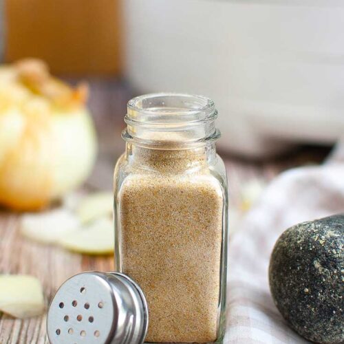 A clear, glass, spice jar sits on a wooden table, filled with homemade onion powder.