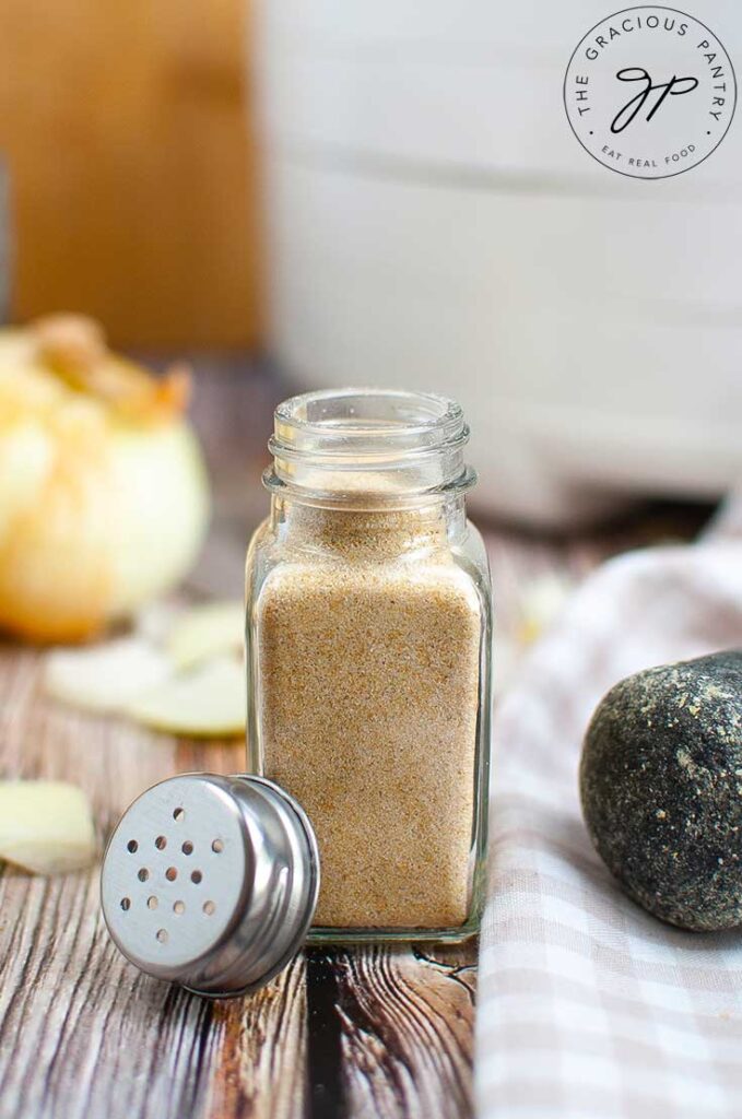 A clear, glass, spice jar sits on a wooden table, filled with homemade onion powder.