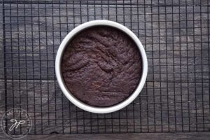 A chocolate cake in a silver cake pan cools on a black wire rack.
