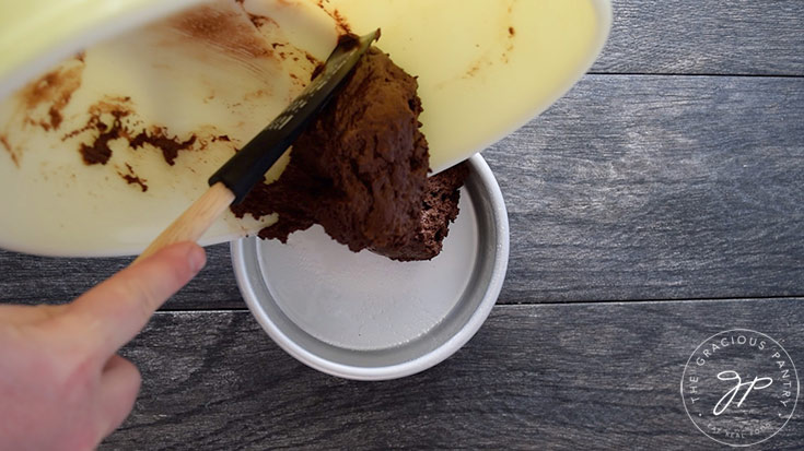 A spatula pushes chocolate batter from a mixing bowl into a small cake pan.