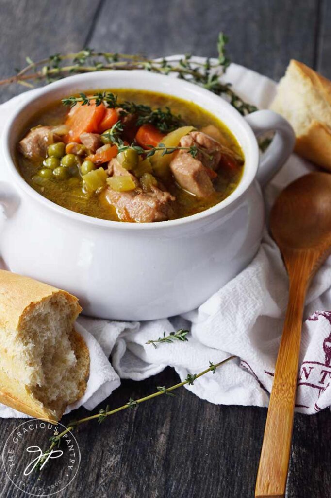 A front view of a white bowl filled with chicken stew and surrounded by pieces of baguette and a wooden spoon.