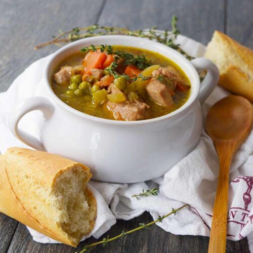 A white bowl with two handles, filled with stew sits on a white towel. Baguette pieces and a wooden spoon surround the bowl.