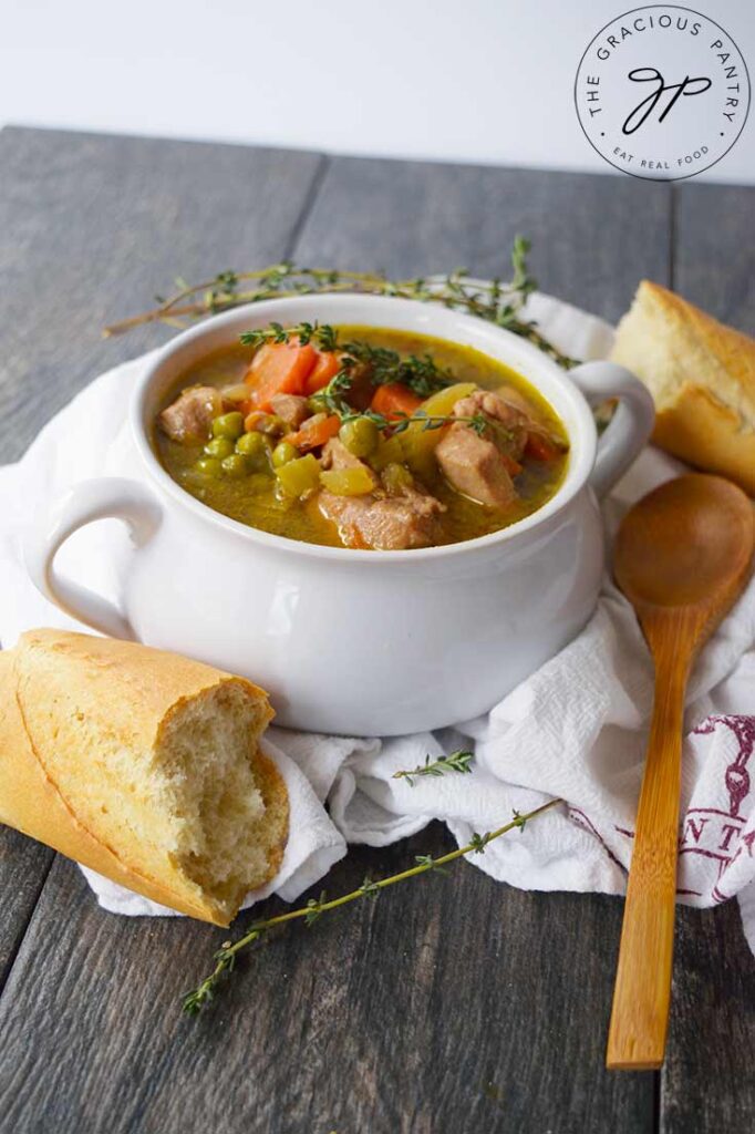 A white bowl with two handles, filled with stew sits on a white towel. Baguette pieces and a wooden spoon surround the bowl.