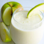 A Tart Green Apple Smoothie in a clear glass. A cut apple sits to the side of the glass.