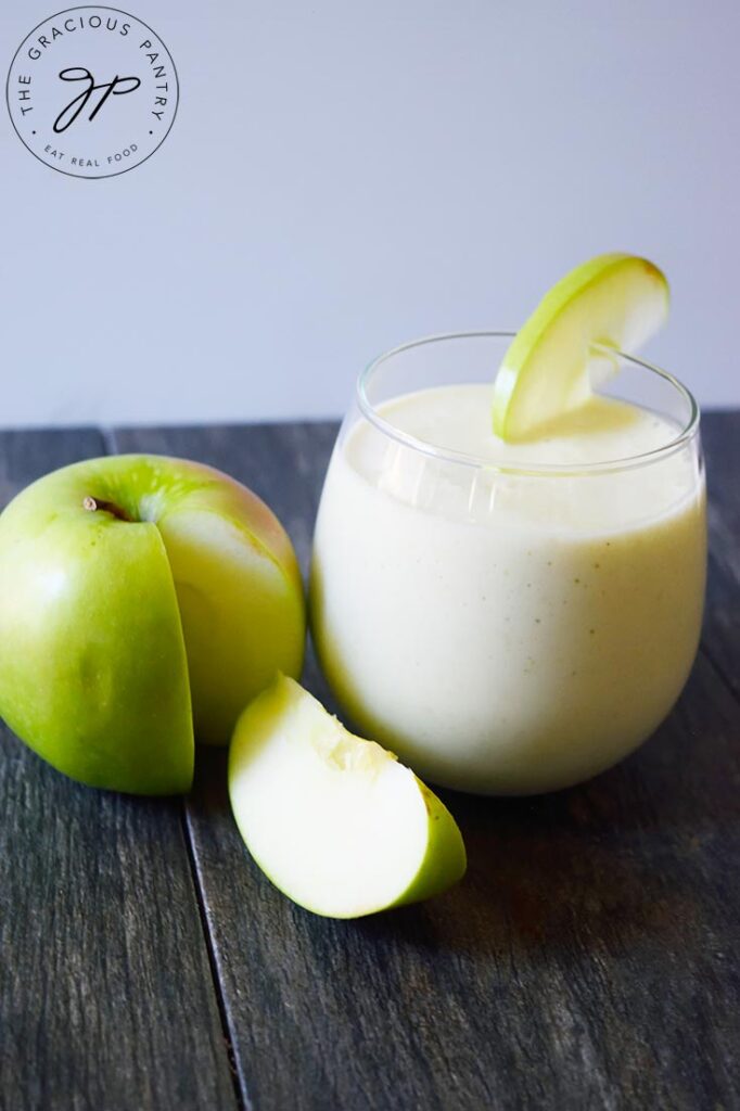 A Tart Green Apple Smoothie in a glass, sitting on a gray counter next to a cut apple.