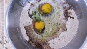 Eggs and liquids added to a flour mixture in a stainless steel mixing bowl.