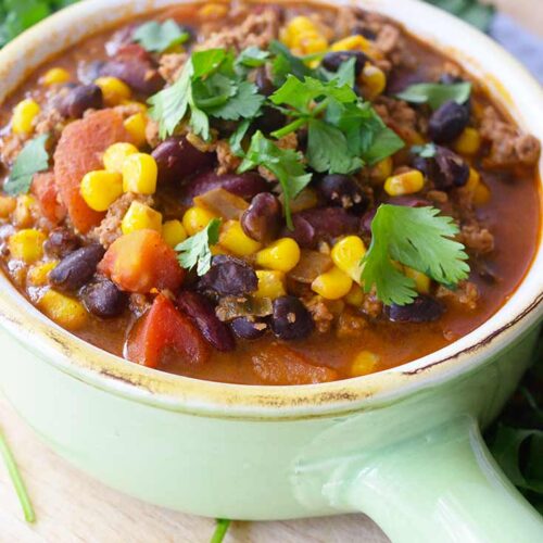 A close up of a light, mint green crock filled with 2 Bean Turkey Chili. Fresh herbs lay just behind the crock.