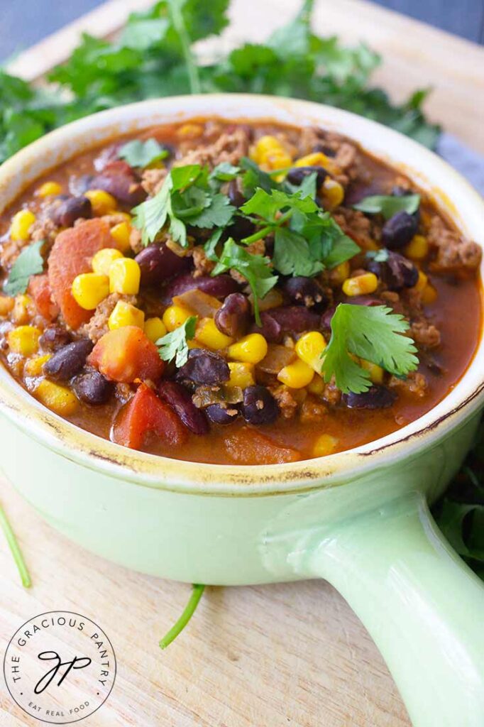 A close up of a light, mint green crock filled with 2 Bean Turkey Chili. Fresh herbs lay just behind the crock.