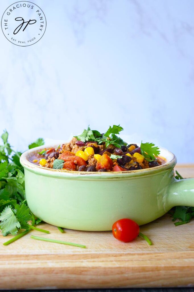 A side view of a light, mint green crock filled with 2 Bean Turkey Chili. The crock sits on a cutting board against a white background.