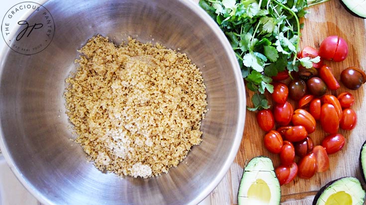 A mixing bowl with quinoa in it. Other recipes ingredients sit on a cutting board to the right of the bowl.