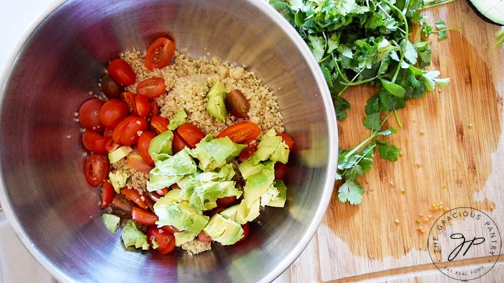 Avocado added to quinoa and sliced grape tomatoes in a mixing bowl.
