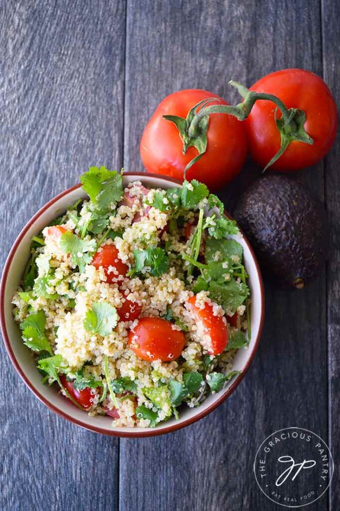 An overhead view looking down into a bowl filled with this Avocado Quinoa Salad. Two tomatoes and an avocado sit next to the bowl.