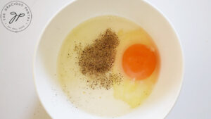 A white mixing bowl with an egg, milk, salt and pepper in it.
