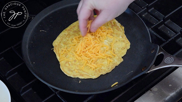 Grated cheese being sprinkled on the egg pancake in a skillet.