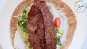 Three strips of bacon laid out over tomatoes, lettuce and mayo on a whole wheat tortilla.