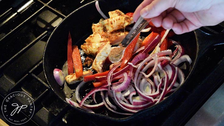 Adding spices to onions, bell peppers and chicken cooking in a cast iron skillet.