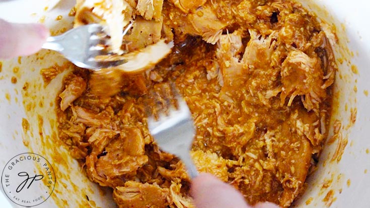 Shredding cooked chicken with forks in a white bowl.