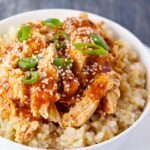 A close up shot of a white bowl filled with Crock Pot Honey Sesame Chicken laying on a bed of brown rice.