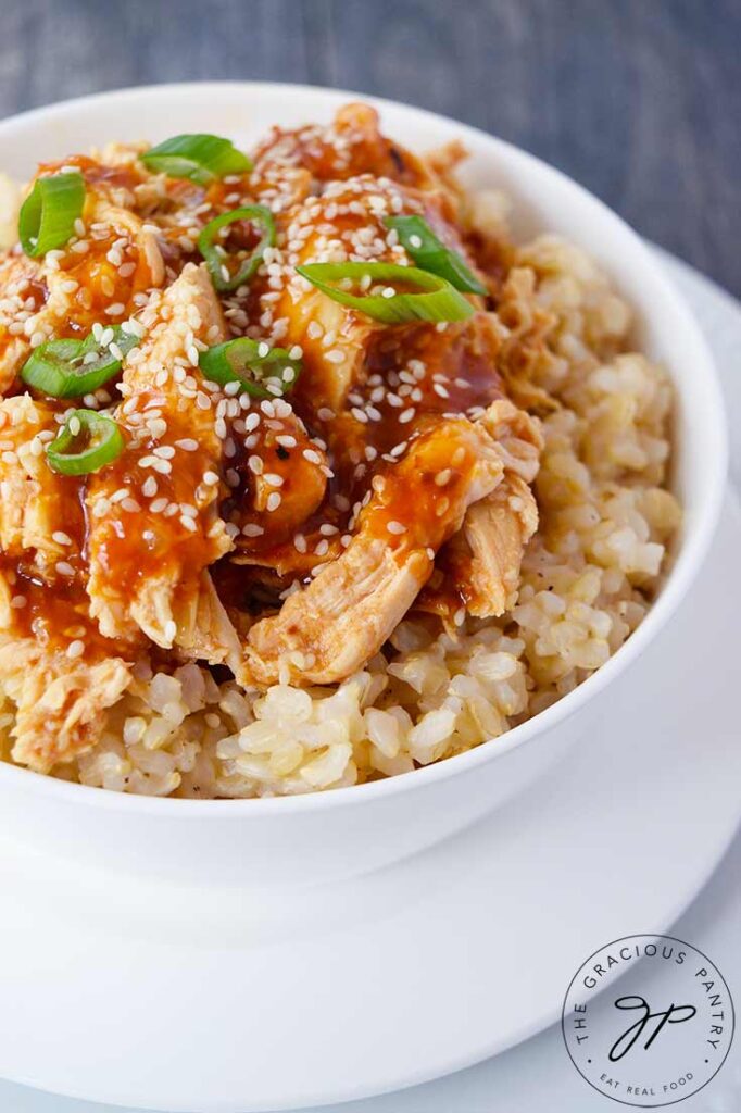 A partial view of a white bowl sitting on white plates, filled with Crock Pot Honey Sesame Chicken and garnished with sesame seeds and slices of green onion.