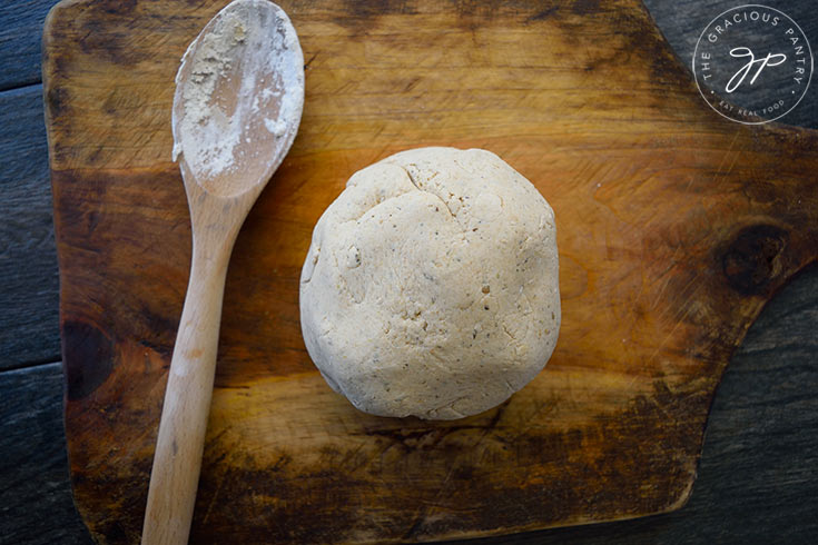 A just finished ball fo pizza dough sits on a wooden cutting board with a used wooden spoon laying next to it with flour and dough on it.