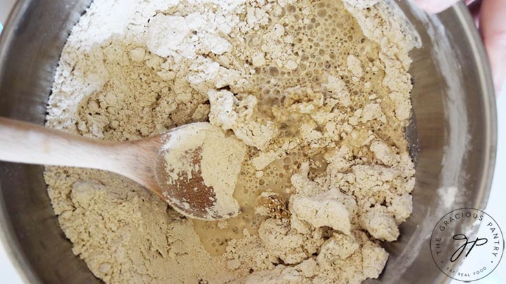 Liquid added to a flour mixture in a mixing bowl. A wooden spoon rests inside the bowl.