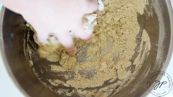 A hand mixing dough in a stainless steel mixing bowl.