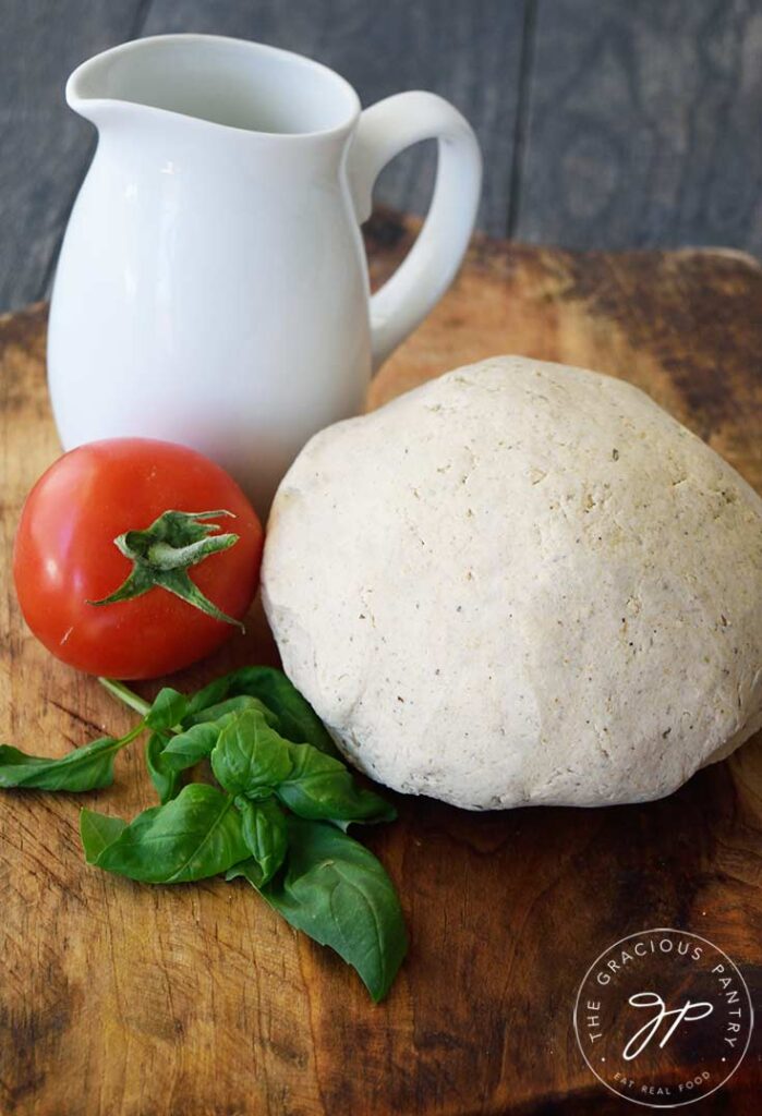 An angled view of a ball of oat flour pizza crust dough sitting on a cutting board next to a white pitcher, a tomato and some fresh basil leaves.