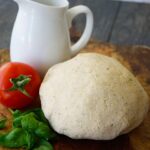 A ball of pizza dough sits on a cutting board next to a white pitcher, a fresh tomato and some fresh basil leaves.