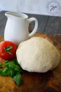 A ball of pizza dough sits on a cutting board next to a white pitcher, a fresh tomato and some fresh basil leaves.