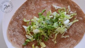 Thinly sliced lettuce laying on a whole wheat tortilla on a white plate.