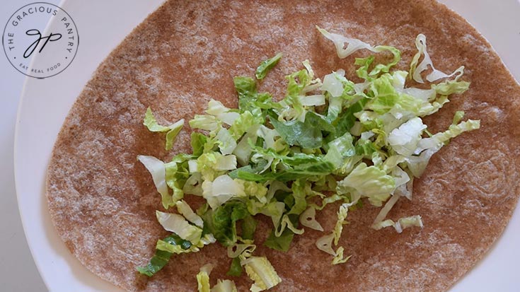 Thinly sliced lettuce laying on a whole wheat tortilla on a white plate.