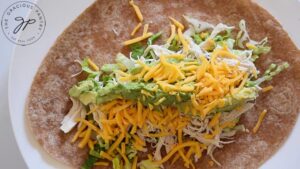 Grated cheddar cheese sprinkled over avocado, shredded chicken, lettuce and a whole wheat tortilla on a white plate.