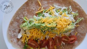 Sliced grape tomatoes laying next to the pile of Ranch Chicken Wrap filling on a whole wheat tortilla.