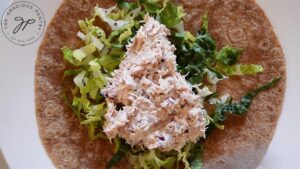 Tuna salad layered on lettuce, sitting on a whole wheat tortilla on a white plate.