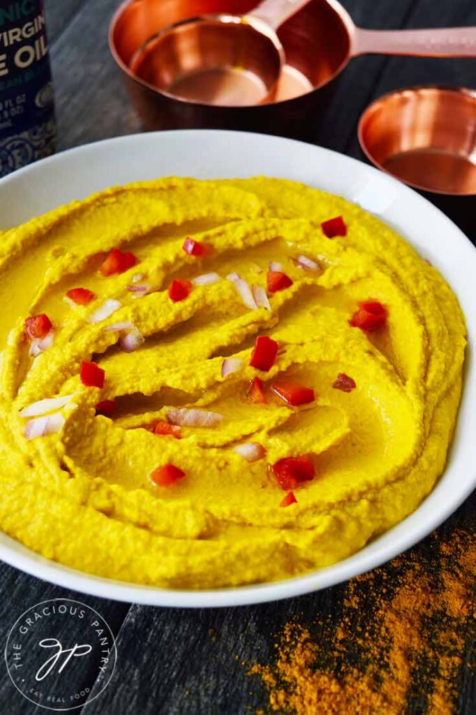 A closeup shot of a white bowl filled with Turmeric Hummus. Copper measuring cups sit on the table behind the bowl.