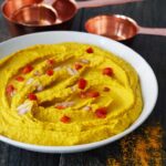 A white bowl filled with Turmeric Hummus and garnished with chopped red peppers and onions.