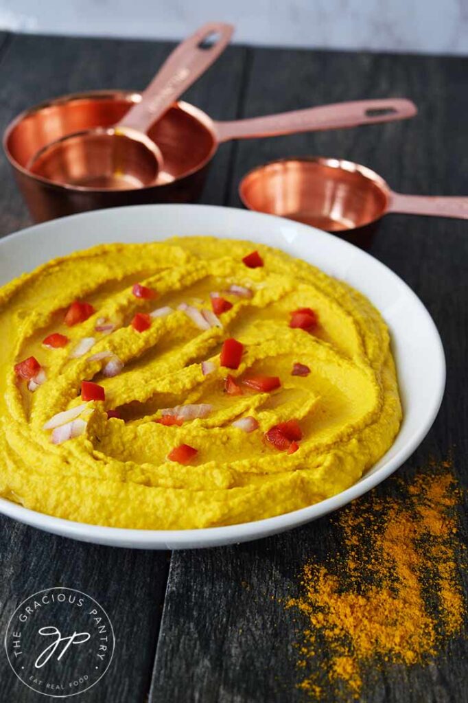 A white bowl filled with Turmeric Hummus and garnished with chopped red peppers and onions.