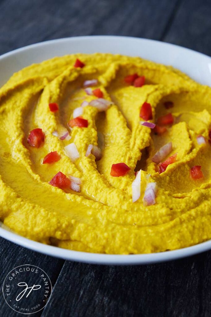 A white bowl filled with Turmeric Hummus and garnished with red peppers and onions.