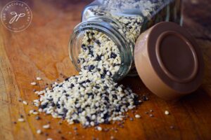 A glass spice jar filled with Everything Bagel Seasoning is tipped over and spilling out onto a wooden surface.