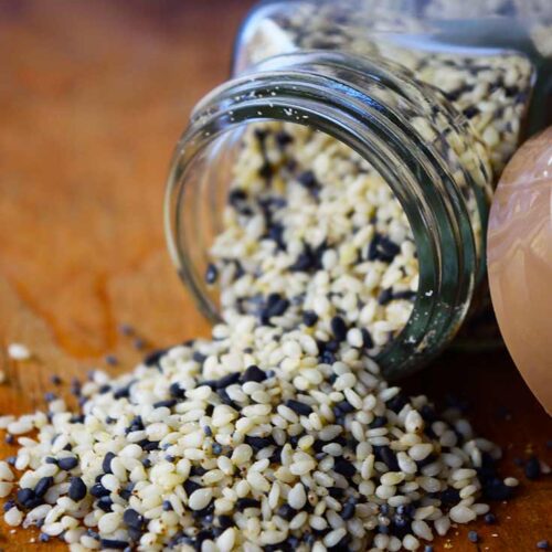 A close up view of a tipped over spice jar with Everything Bagel Seasoning spilling out onto a wooden surface.