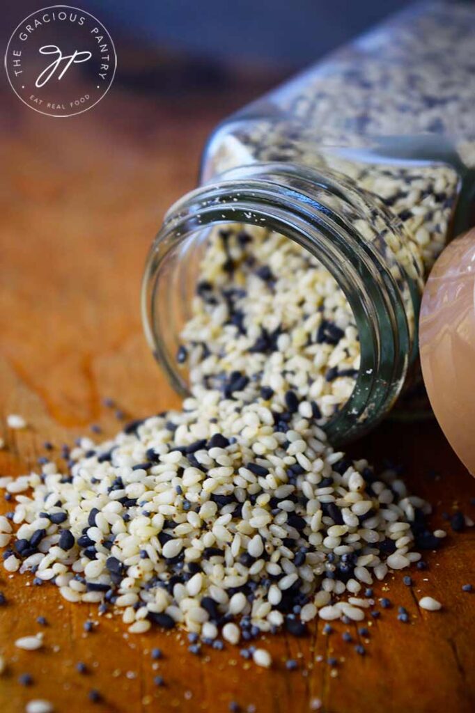 A close up view of a tipped over spice jar with Everything Bagel Seasoning spilling out onto a wooden surface.