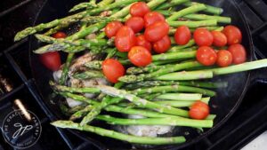 Grape tomatoes added to asparagus and chicken thighs in a cast iron skillet.
