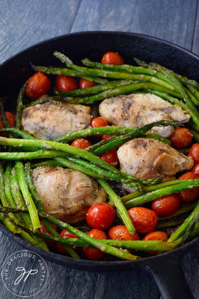 A black, cast iron skillet holds chicken thighs, asparagus and grape tomatoes.