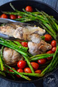 A pair of tongs hold a chicken thigh for the camera, over a cast iron skillet filled with oven-baked chicken thighs, asparagus and grape tomatoes.