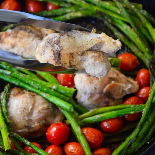 A pair of tongs hold a chicken thigh for the camera, over a cast iron skillet filled with oven-baked chicken thighs, asparagus and grape tomatoes.