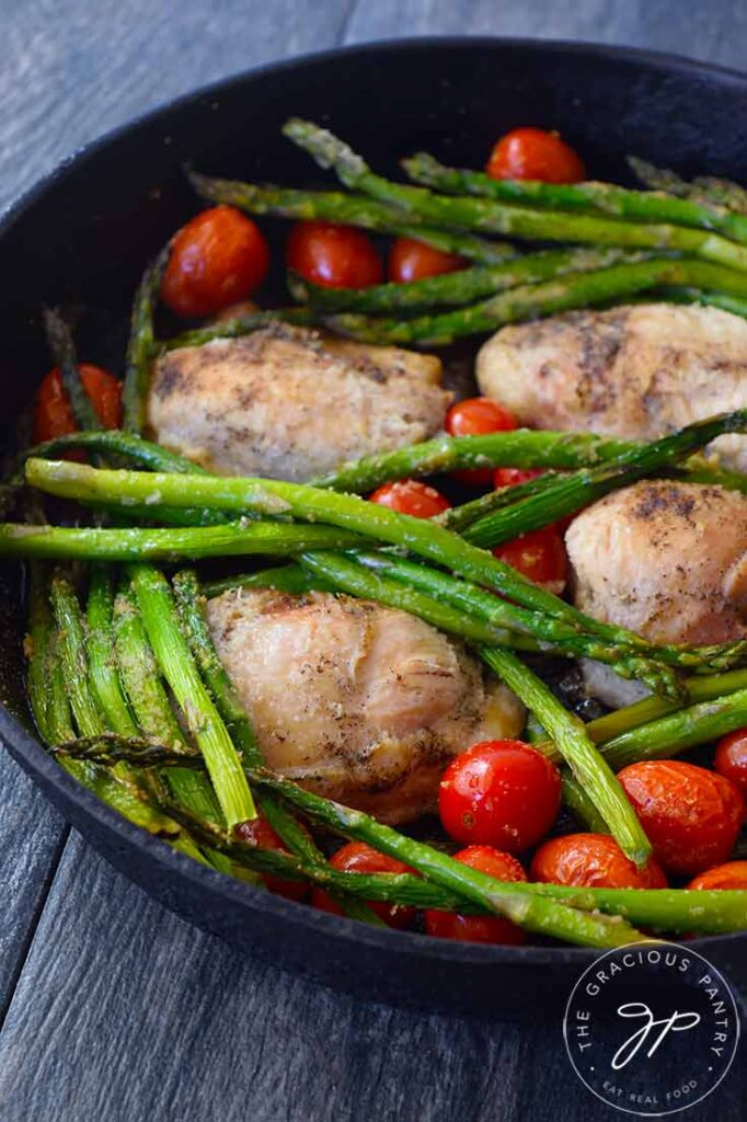 A side view of Oven-Baked Chicken Thighs in a cast iron skillet with asparagus and grape tomatoes.