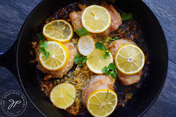 An overhead view of a black skillet filled with just baked garlic chicken thighs. Lemon slices lay on top of each chicken thigh.