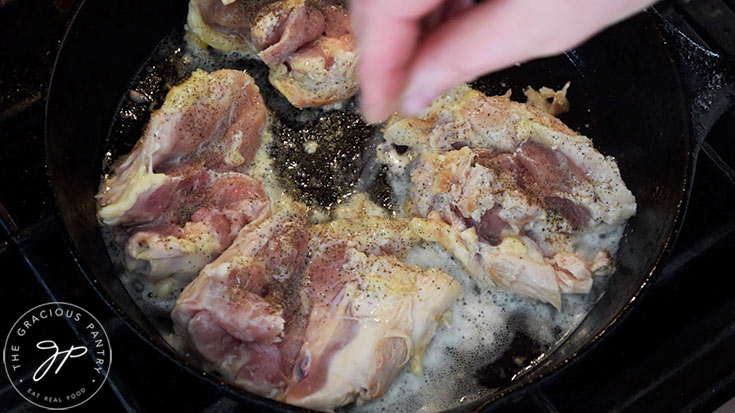 Seasonings being sprinkled over chicken thighs as they brown in a skillet.