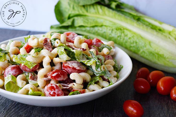 BLT Pasta Salad in a white bowl with a head of lettuce and some grape tomatoes sitting behind the bowl.